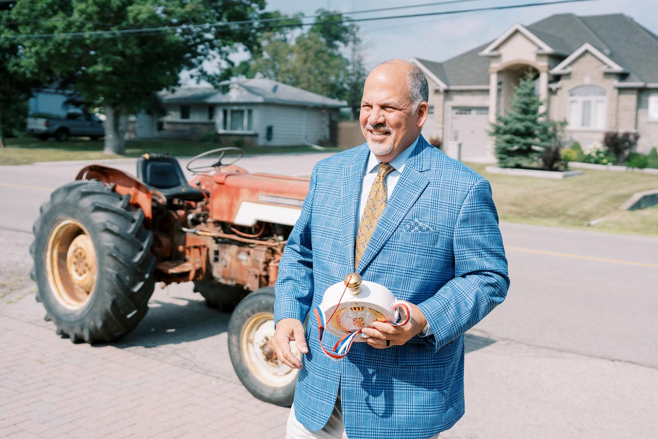Man in a blue checkered suit smiling while holding a white trophy with ribbons, standing next to an old red tractor on a suburban street.