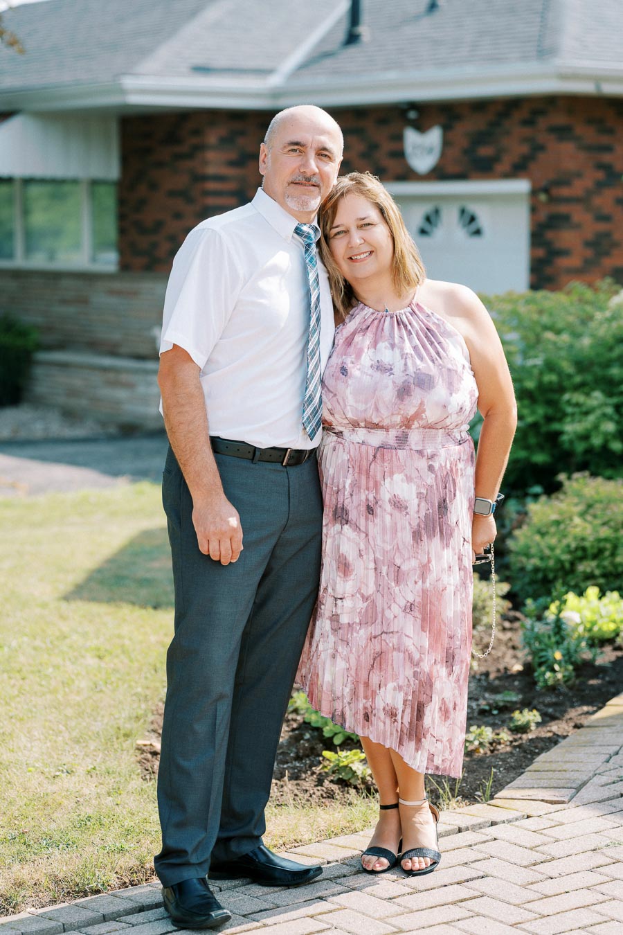 A couple stands smiling in front of a brick house, surrounded by greenery. The man wears a white shirt and tie, while the woman is dressed in a floral pink dress. The scene is cheerful and sunny, ideal for a family or real estate photo.