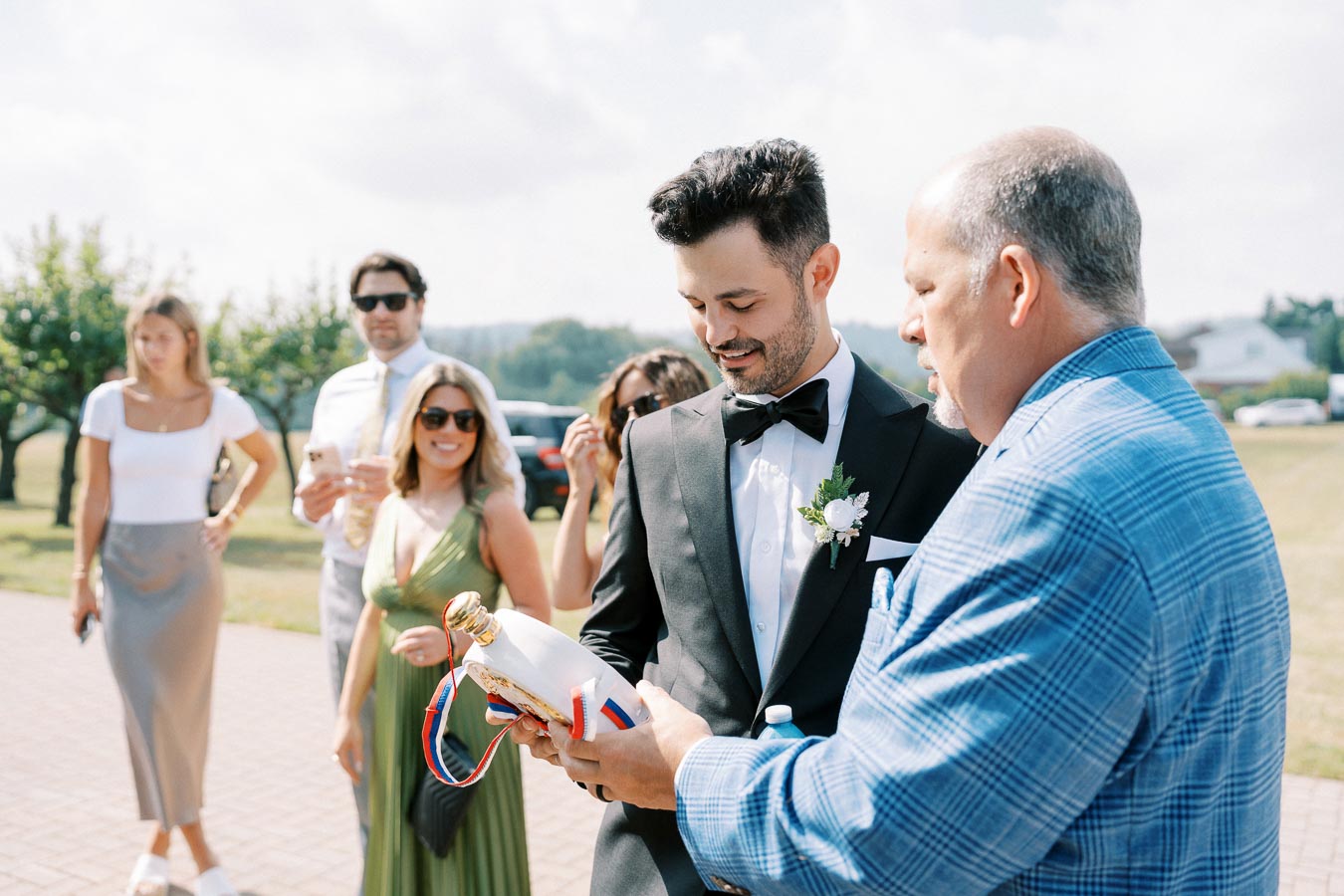 A group of well-dressed wedding guests gathers outdoors on a sunny day. The groom, wearing a black tuxedo with a white boutonnière, smiles as he receives a gift bottle from another guest in a blue suit. Other attendees, including a woman in a green dress and people in formal attire, look on.