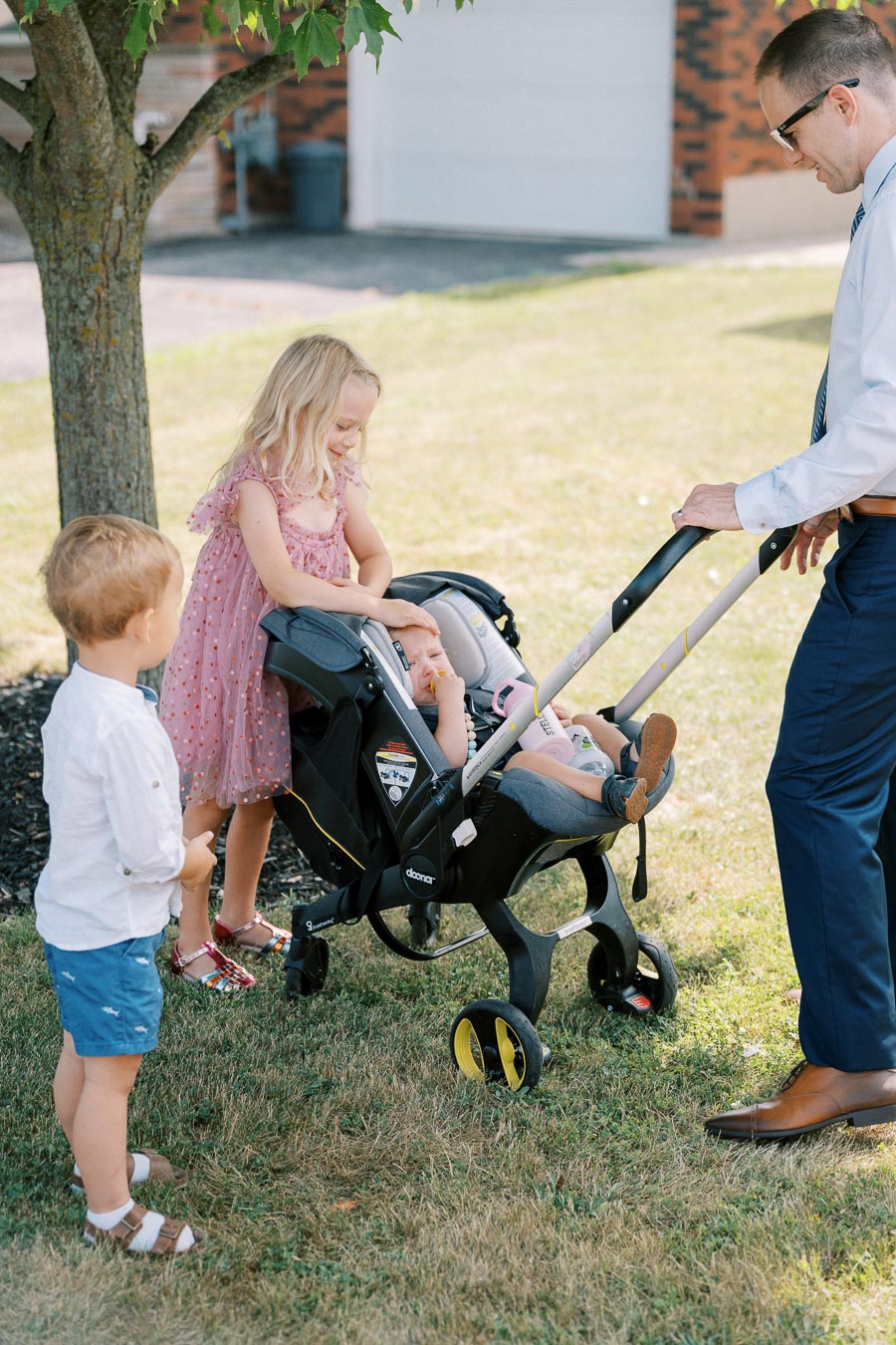 A man in a suit pushes a stroller with a baby, while two young children stand nearby on a grassy lawn under a tree. The scene captures a sunny, outdoor family moment in a suburban setting.