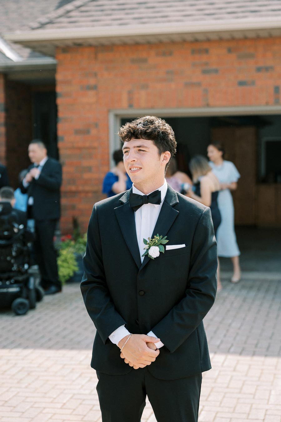 A young man in a black tuxedo and bow tie stands outside a brick building. He has a white boutonniere pinned to his jacket. Other people are dressed in formal attire in the background, socializing outdoors on a sunny day.