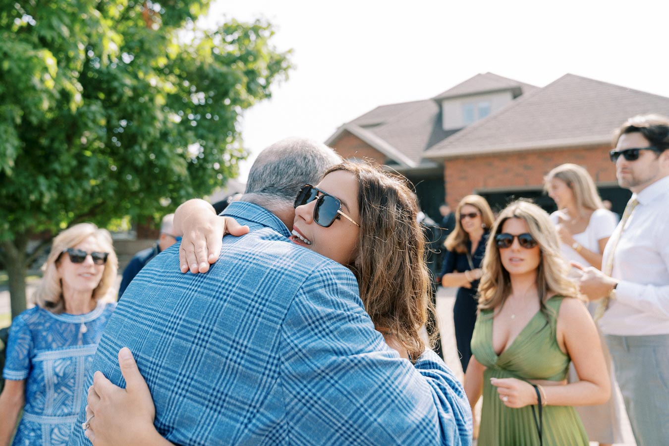 A woman embraces a man wearing a blue checked jacket outdoors, surrounded by a group of people at a festive gathering or celebration. Everyone is dressed in summer attire, wearing sunglasses, and standing near a house with a lush tree in the background.