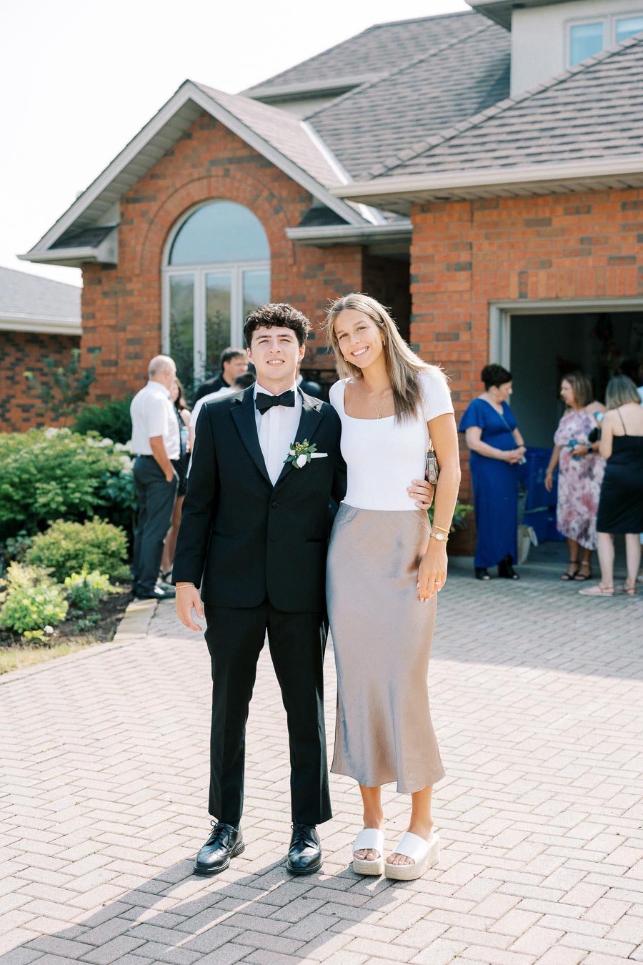 A young couple dressed for a formal event stands smiling in front of a brick house. The man is wearing a black tuxedo with a bow tie while the woman is in a white top and a shiny silver skirt. There are other guests socializing in the background.