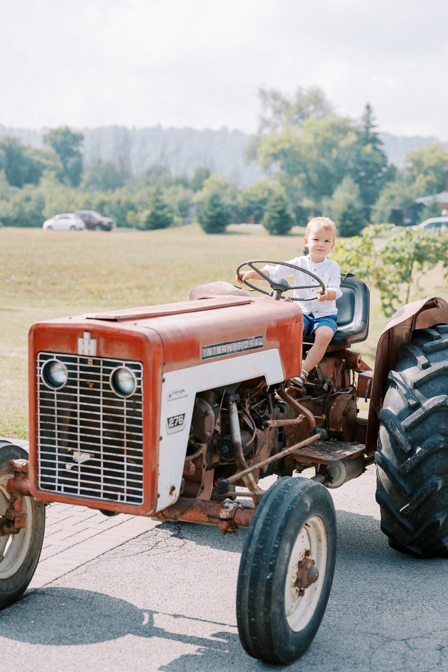 Child sitting on a vintage red tractor in a rural field setting, surrounded by greenery and distant trees under a cloudy sky.