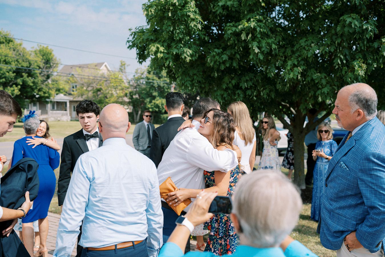 A group of people at a social gathering outdoors, with some individuals hugging and others conversing, under a tree on a sunny day.