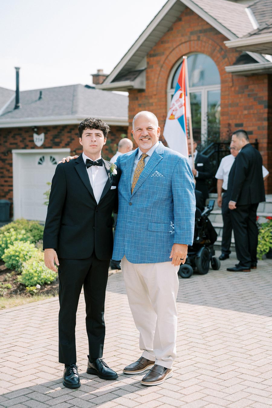 Father and son dressed in formal attire standing outside a brick house, posing for a photo on a sunny day. The young man is wearing a black suit with a bow tie, while the older man wears a blue plaid blazer. A flag is visible in the background.