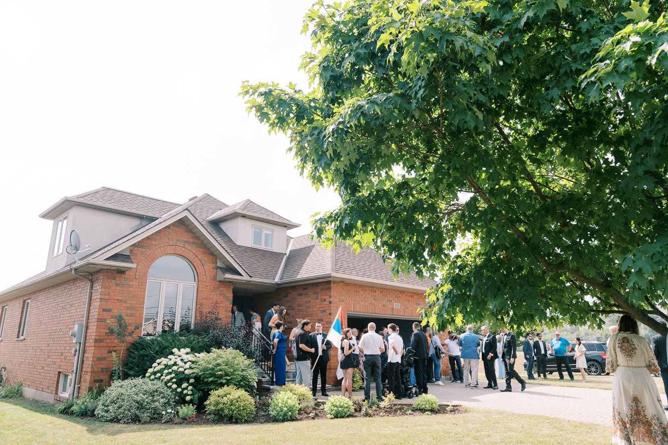 Group of people dressed in formal attire gathering outside a red brick suburban house, with a large green tree in the foreground, during a sunny day event.