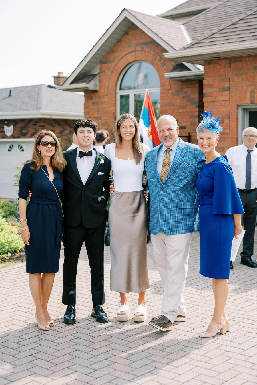 A group of five people dressed in formal attire smiling outside a brick house on a sunny day.