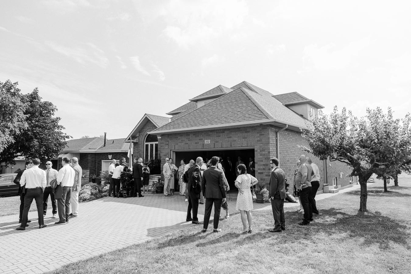 Black and white image of a group of people gathered outside a brick house on a sunny day, with trees and a large driveway visible.