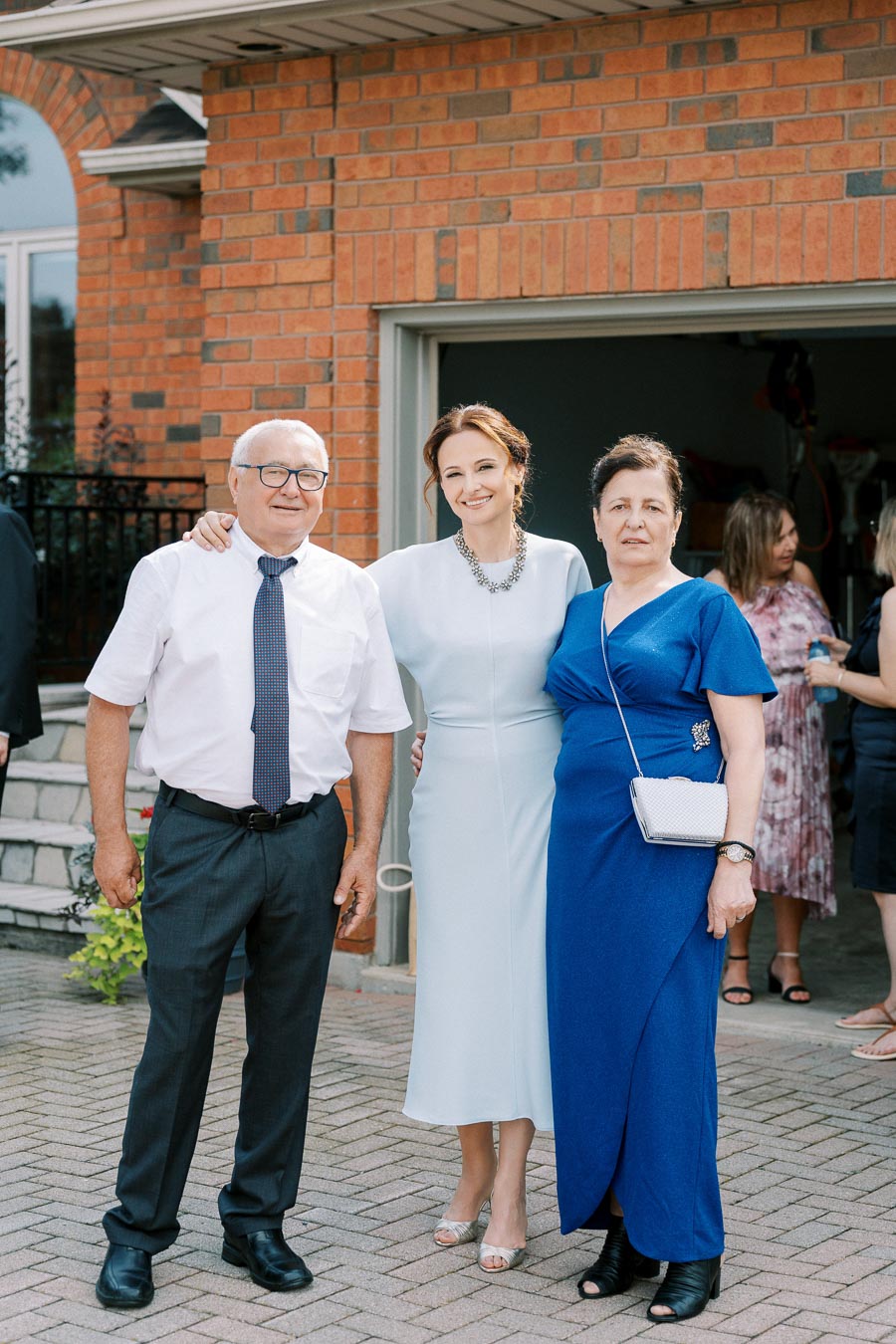 A smiling woman in a light blue dress stands between two older individuals, one wearing a white shirt with a tie and the other in a vibrant blue dress, posing together in front of a brick house.