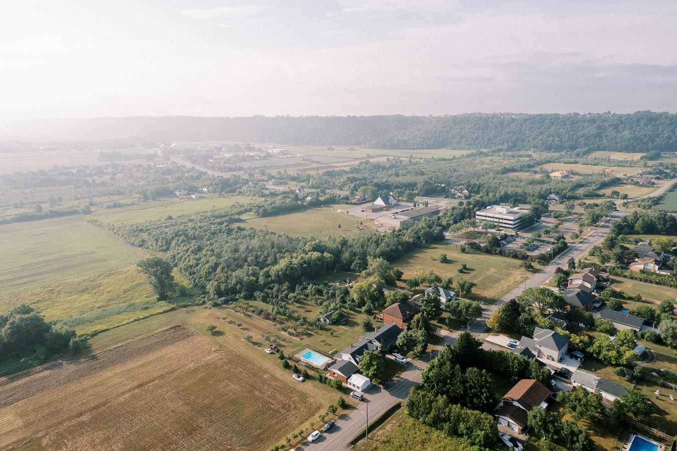 Aerial view of a rural landscape showcasing farmland, residential houses, and surrounding greenery under a clear sky.