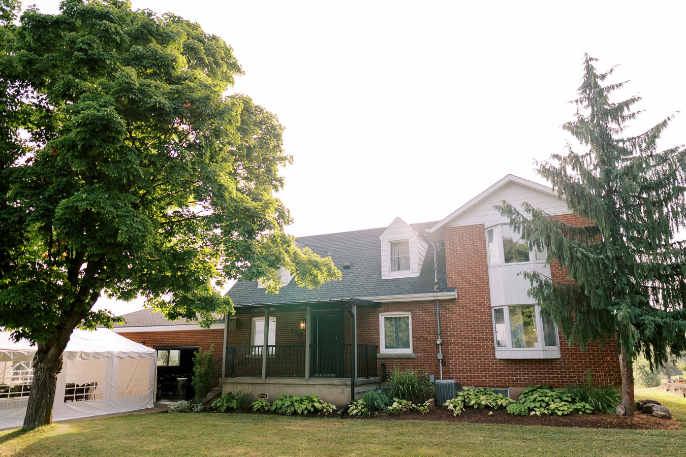 Charming red-brick house with a sloped roof surrounded by lush greenery and trees, featuring a large tent on the left side. A well-maintained lawn and garden add to the inviting appearance of the residential property, captured under bright daylight.
