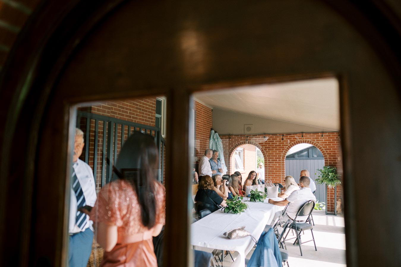 A group of people having a conversation around a table on a covered patio with arched brick openings.