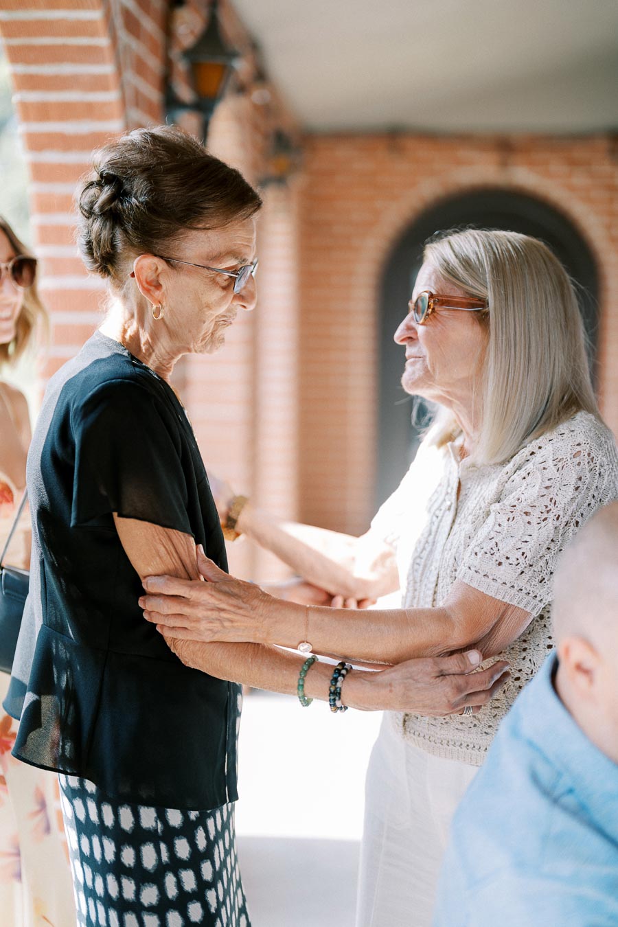 Two elderly women warmly embrace and smile in a sunlit outdoor setting with brick arches in the background.