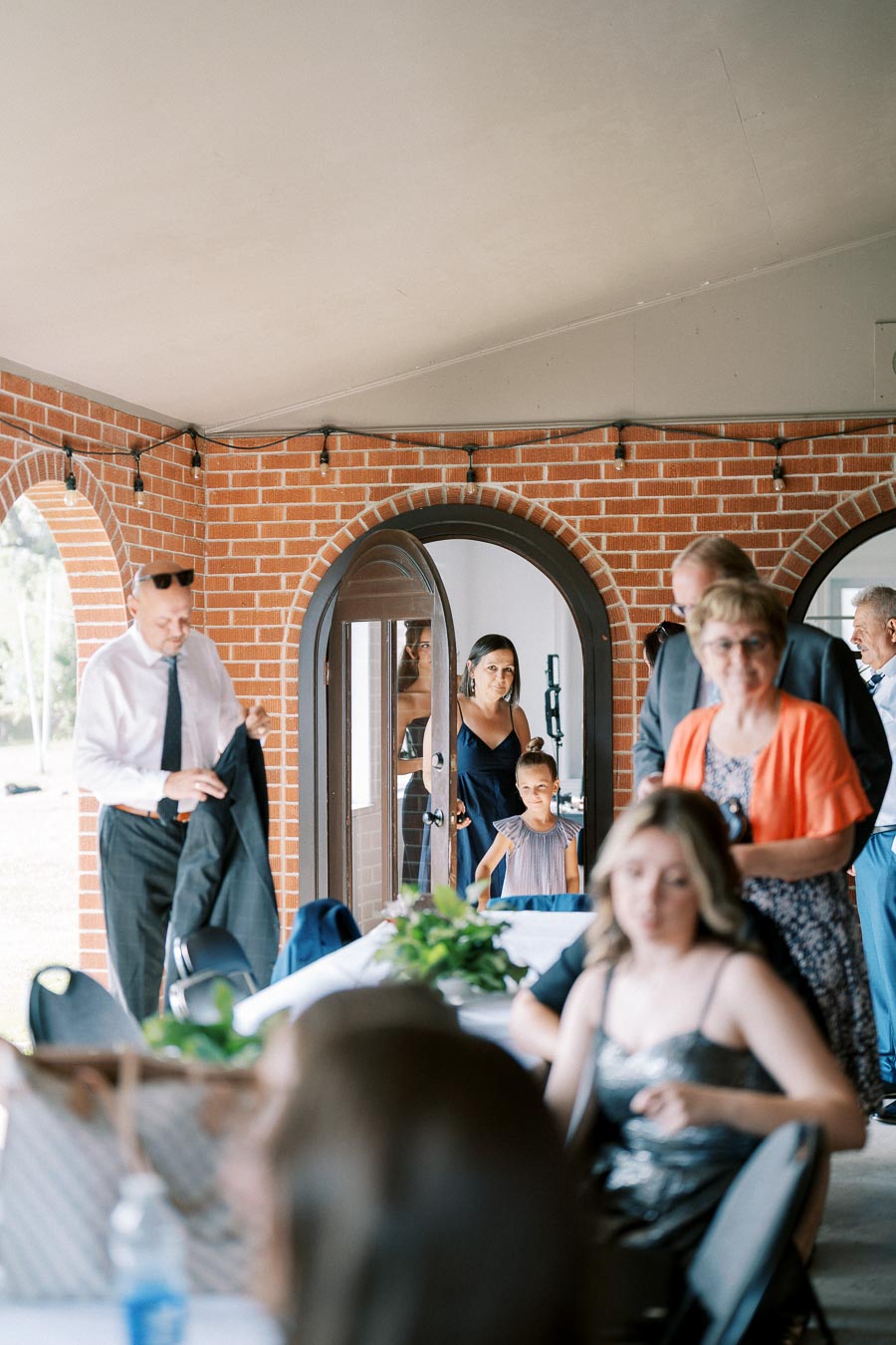 A group of people attending a formal event in a room with brick arches and string lights, featuring a man in a suit and a woman and child entering through an arched doorway.