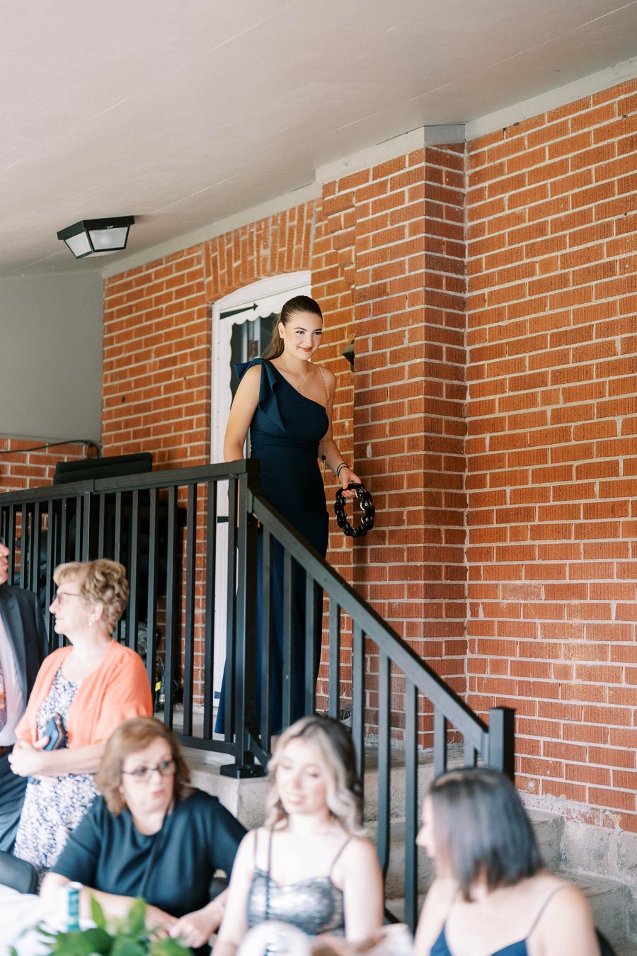 Young woman in elegant dress descending stairs at an outdoor event, holding a tambourine. She is smiling, with attendees sitting at tables below. The setting features a red brick wall, creating a festive atmosphere.