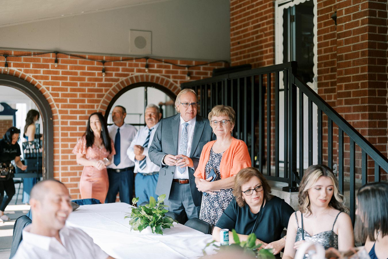 A group of people smiling and socializing at a festive indoor event near a brick wall with a staircase, featuring a table adorned with greenery in the foreground.