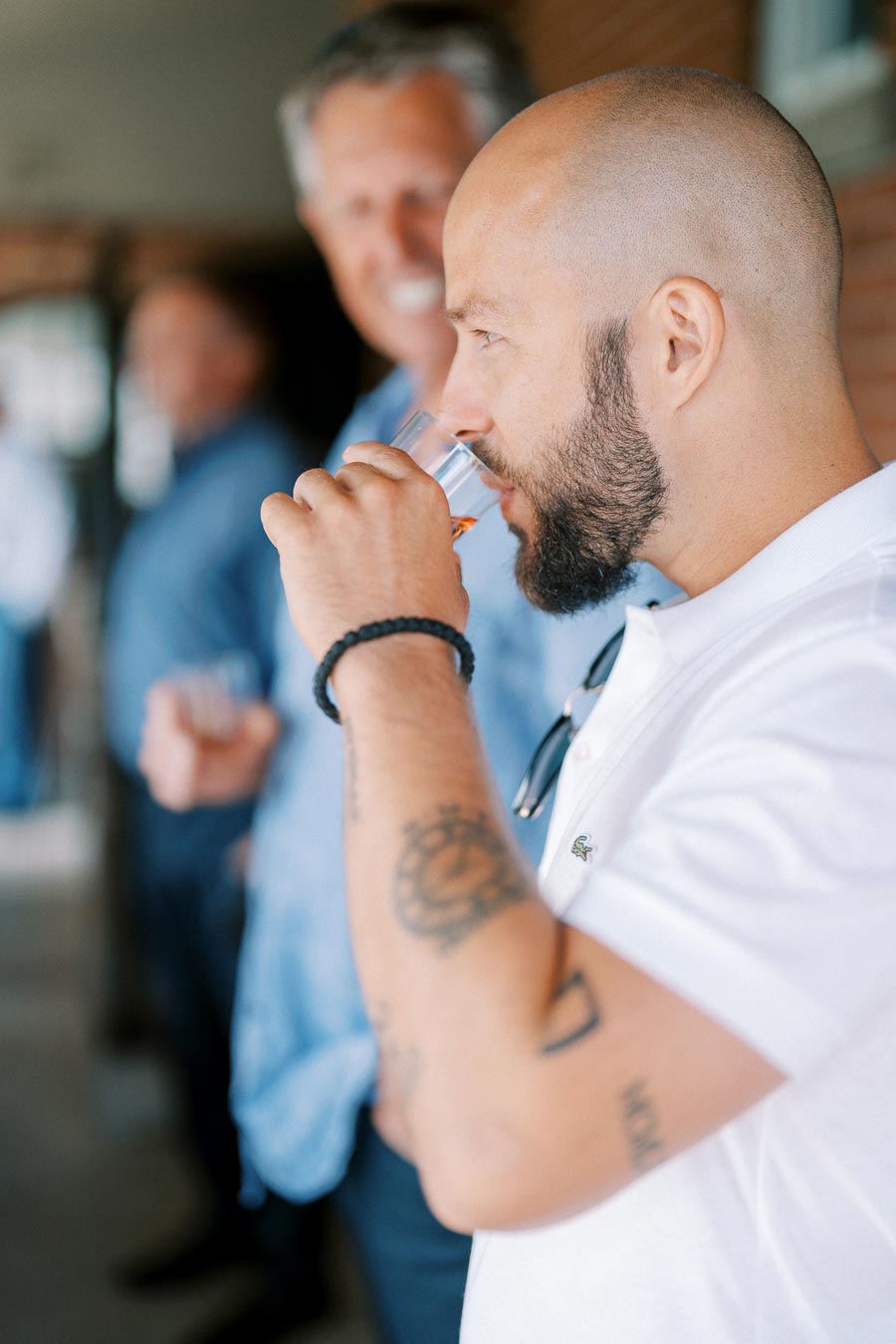 Man with tattoos drinking from a glass at a social gathering, wearing a white shirt, in casual indoor setting with blurred background.