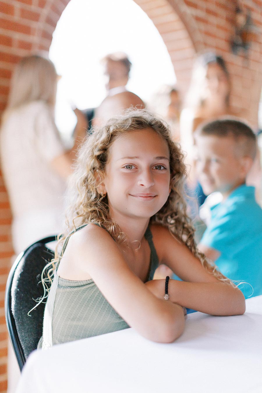 Young girl with curly hair smiling at a table during a social event, with blurred people in the background and brick archways visible.