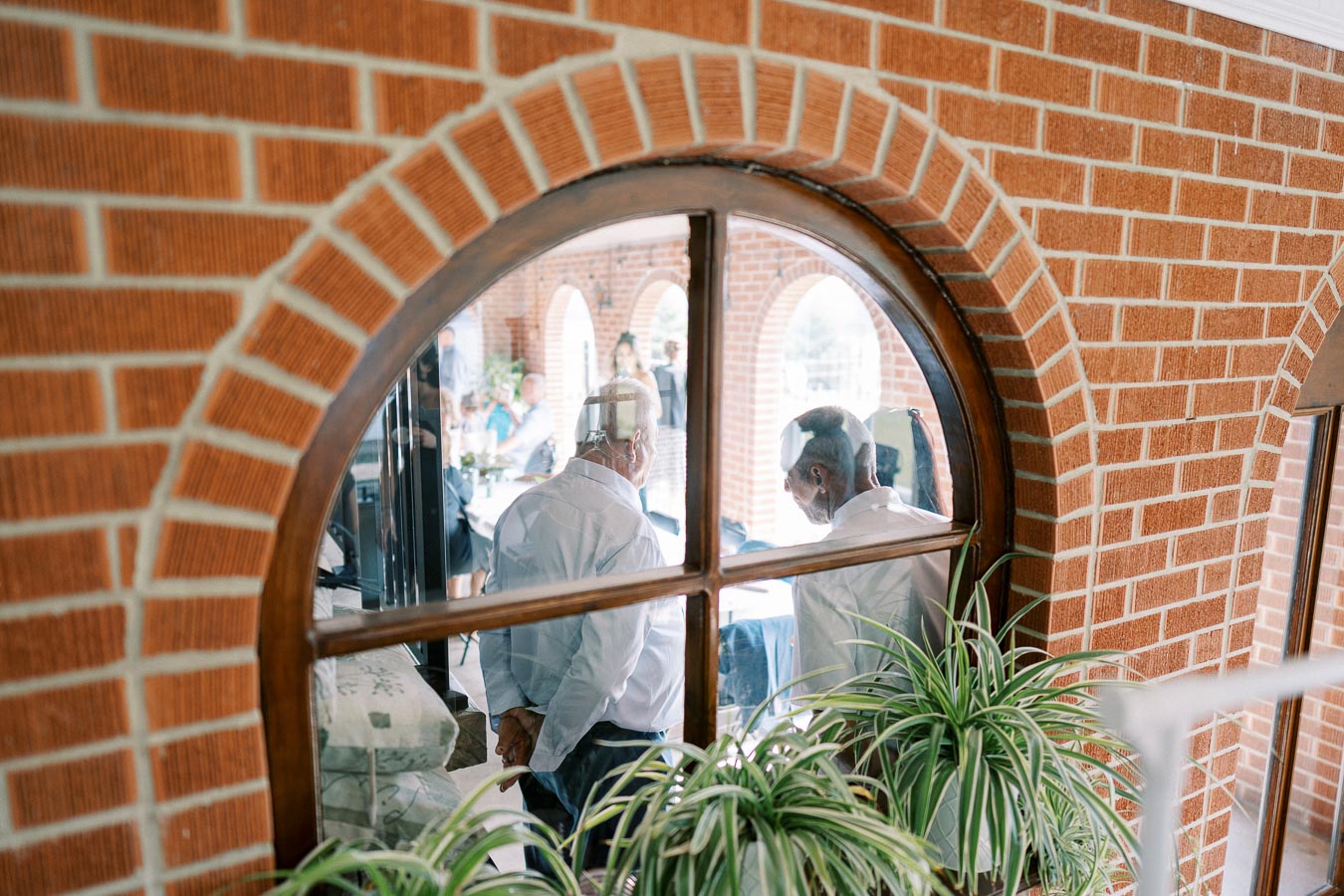 Two people seen through a rounded brick archway window, engaged in conversation, with a decorative plant in the foreground, hinting at a casual gathering or social event indoors.