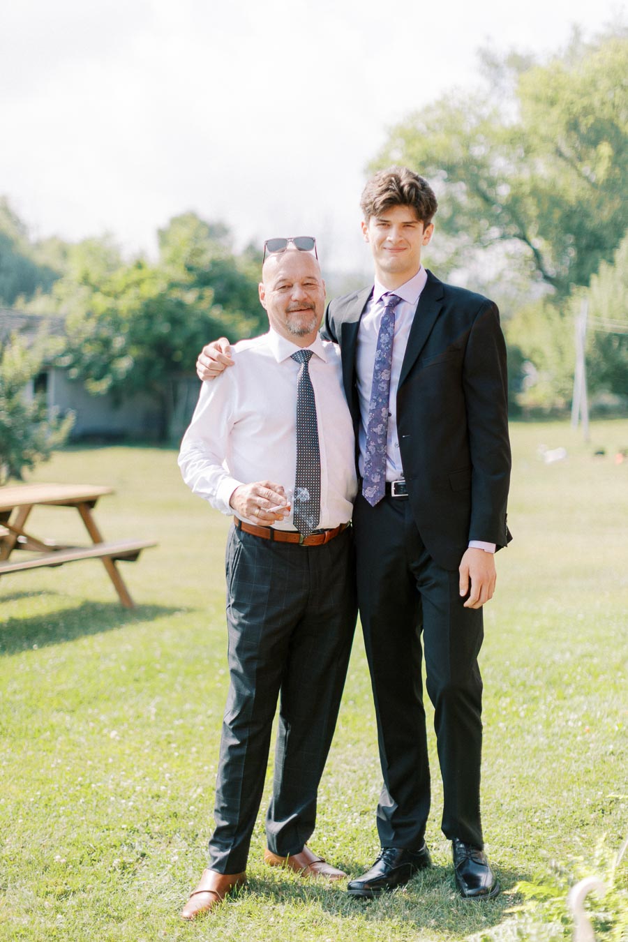 Two people dressed in formal attire standing outdoors on a sunny day, with grass and trees in the background.