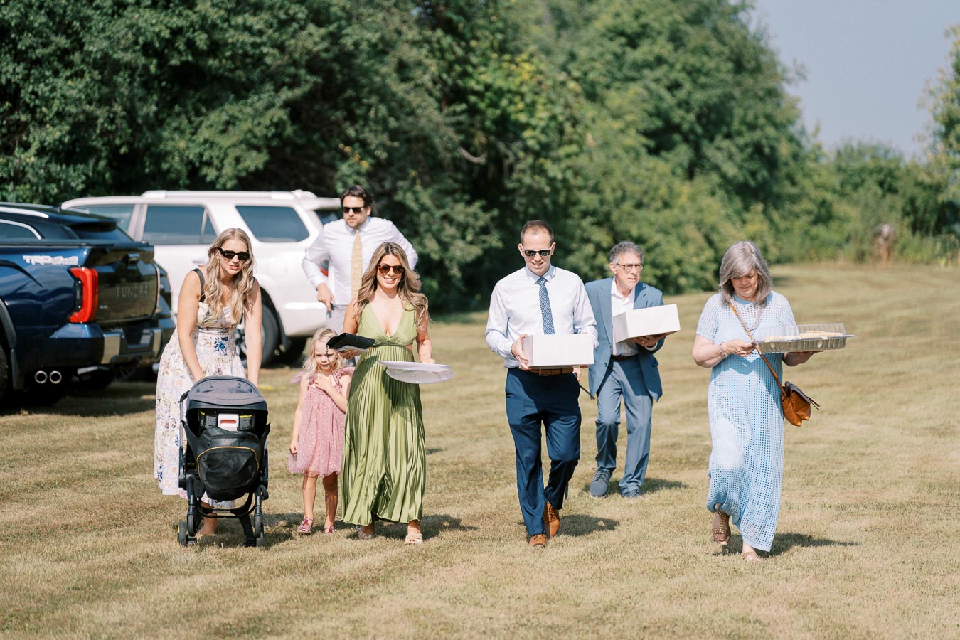 A group of people casually dressed in formal attire walk across a grassy area carrying trays and boxes, with parked cars and lush greenery in the background. The sunny setting suggests they might be heading to a picnic or outdoor gathering.