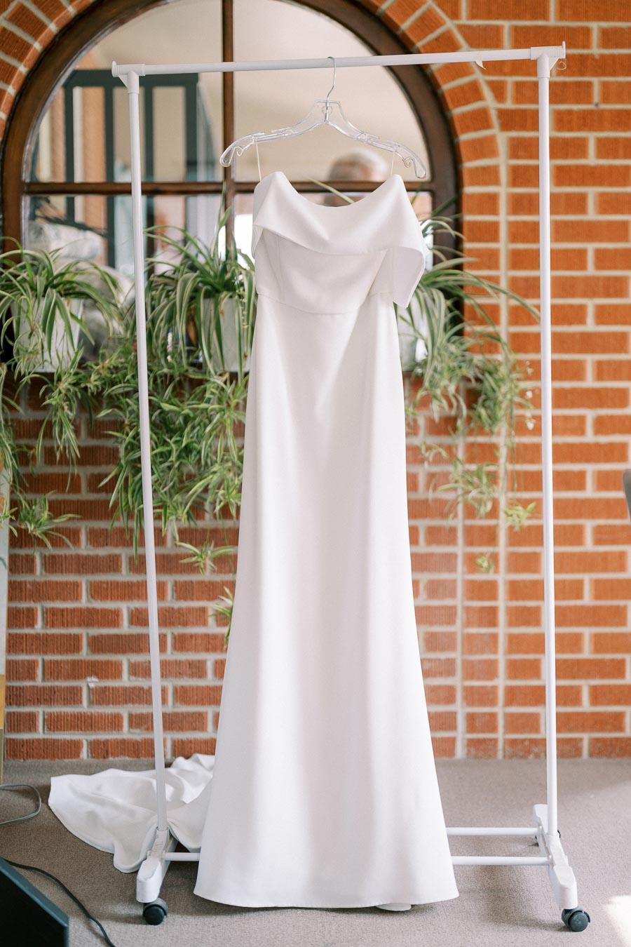 Elegant white strapless wedding dress hanging on a rack in front of a brick wall background, surrounded by green plants, ready for a beautiful ceremony.