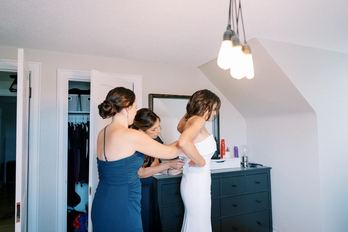 Two bridesmaids assist a bride in a white dress, helping her with final adjustments in a well-lit room with modern decor, preparing for the wedding ceremony.