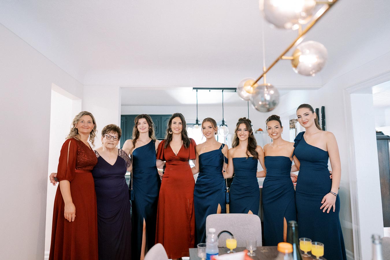 Group of women in elegant blue and red dresses posing together in a well-lit room with a modern light fixture.