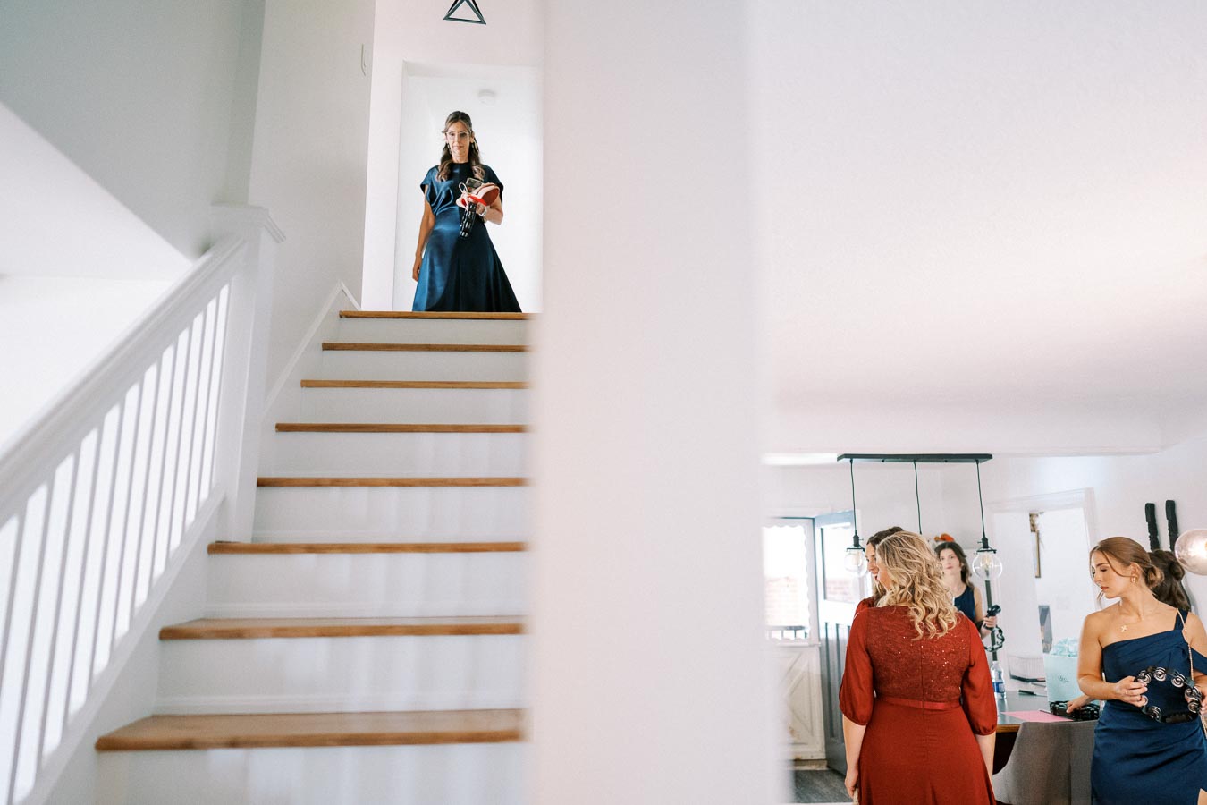 Woman in a blue dress descends a staircase while others converse in the living room below, showcasing a modern home interior with a bright, open layout.