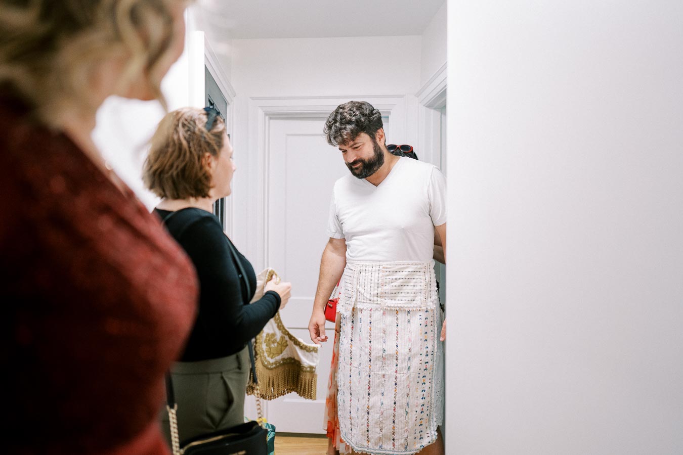 A man wearing a handmade apron smiles while standing in a hallway with two women, who appear to be engaged in conversation. The setting is an indoor space with white walls and a wooden floor, suggesting a casual and friendly atmosphere.