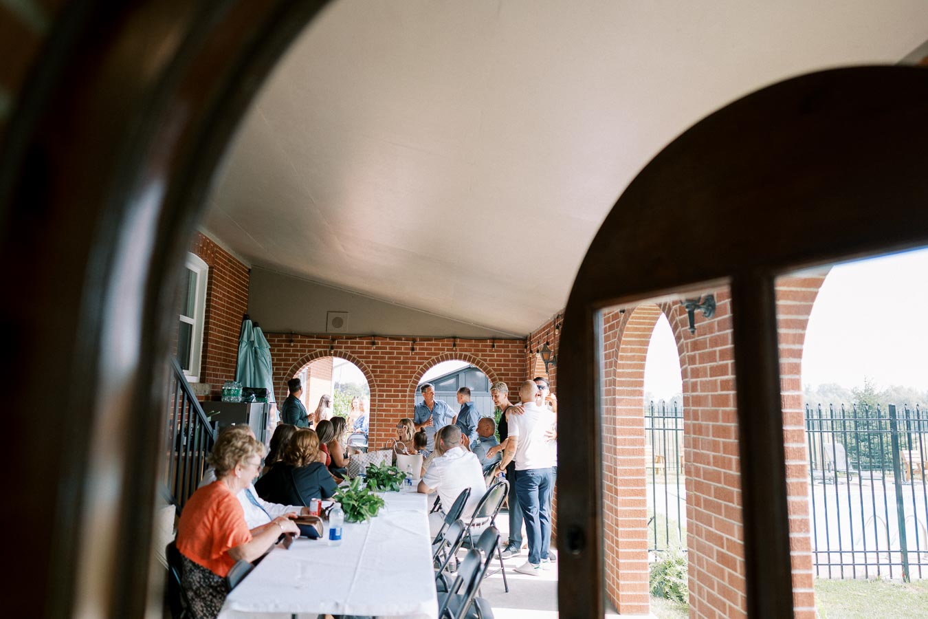 Group of people gathered on a covered porch with arched brick openings, sitting and chatting around a long table with a white tablecloth, during a daytime outdoor event.