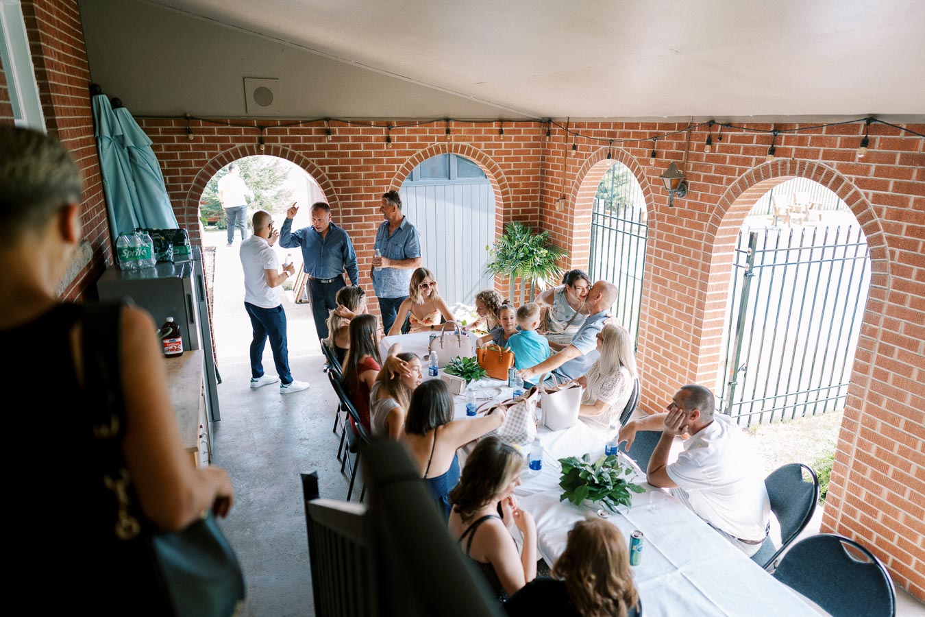 Outdoor family gathering under a brick archway, featuring a group of people sitting around a long table with a white tablecloth, engaged in conversation and enjoying the event.