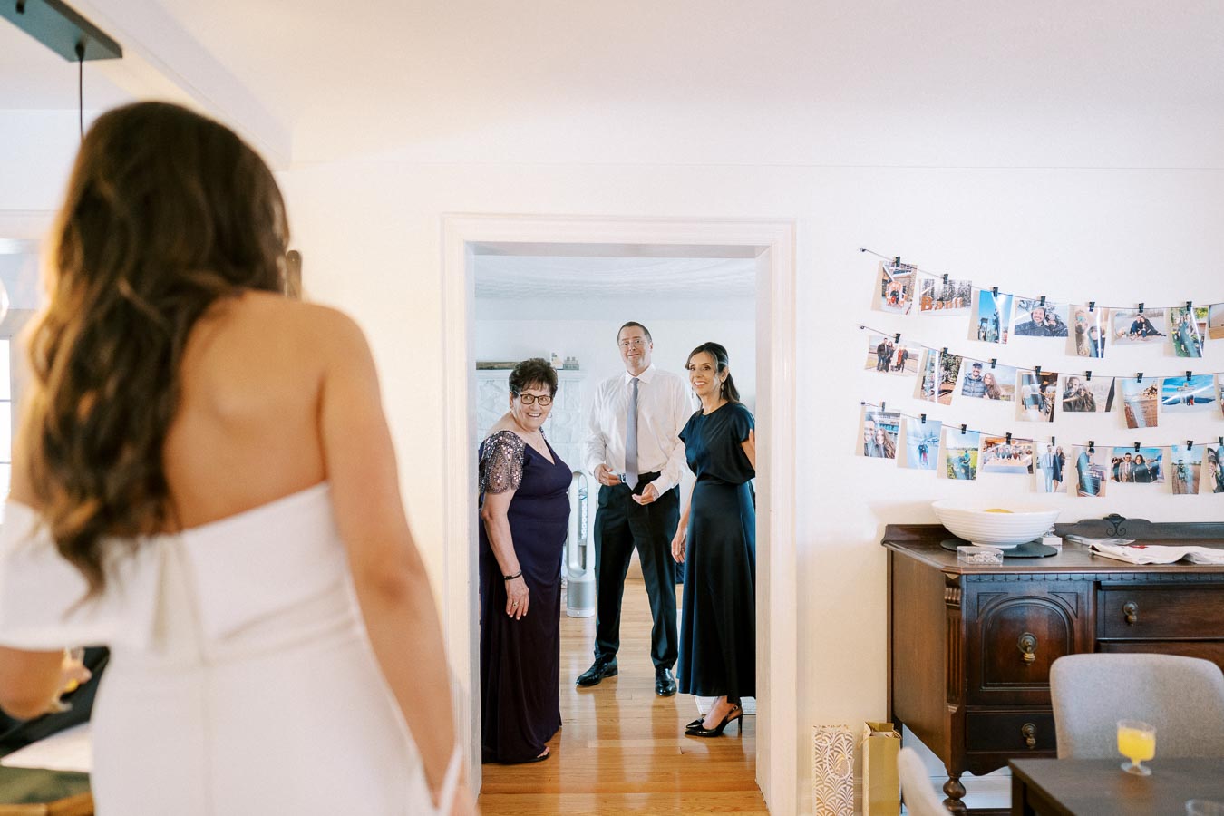 A bride in a white wedding dress is seen from behind, facing three family members standing in a doorway. They appear excited and smiling. The room has a line of photos on the wall and a wooden side table with a bowl and decorative items. Perfect candid moment before a wedding ceremony.