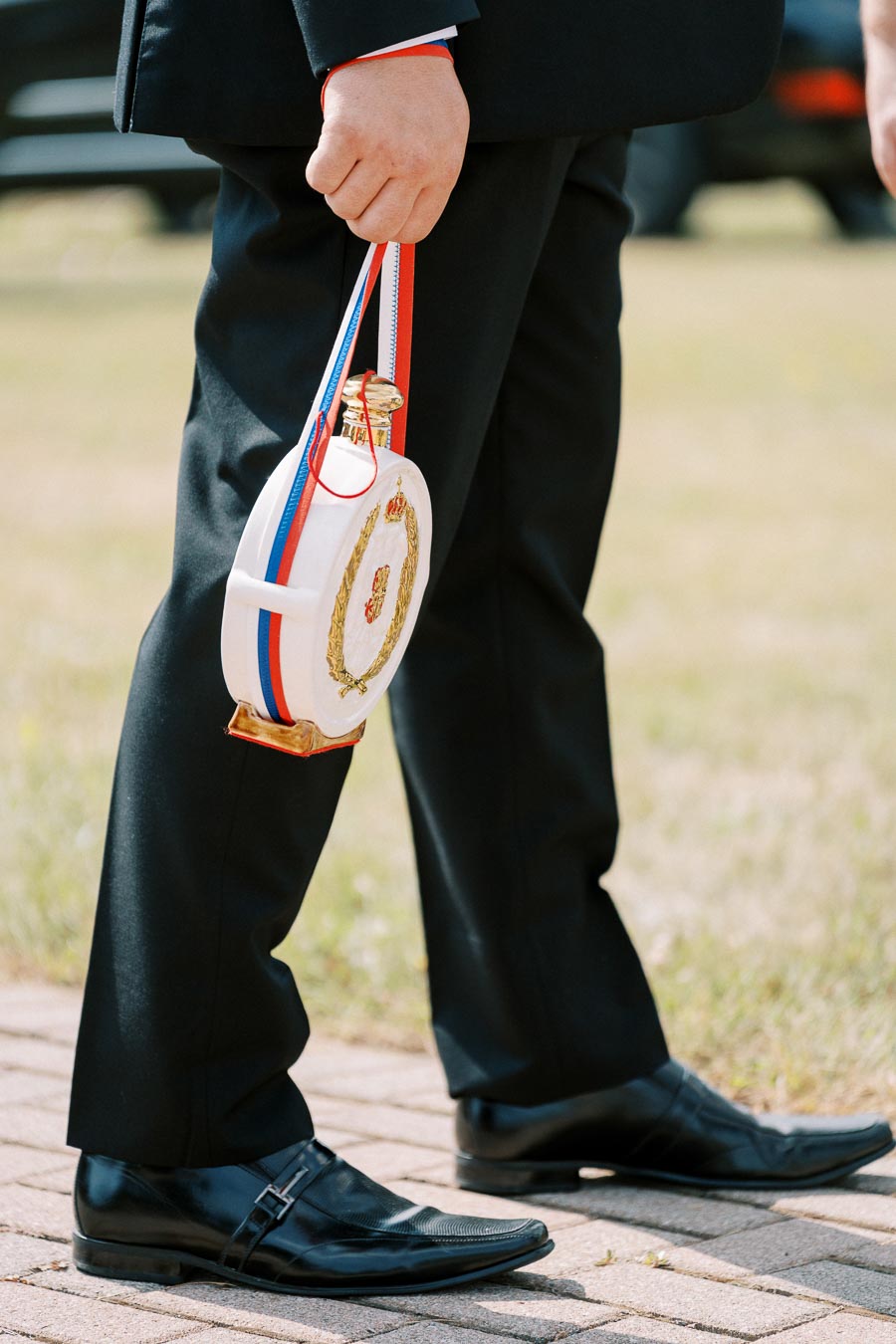 A person in a formal black suit and shoes holds a ceremonial flask decorated with red, white, and blue accents while walking on a path.