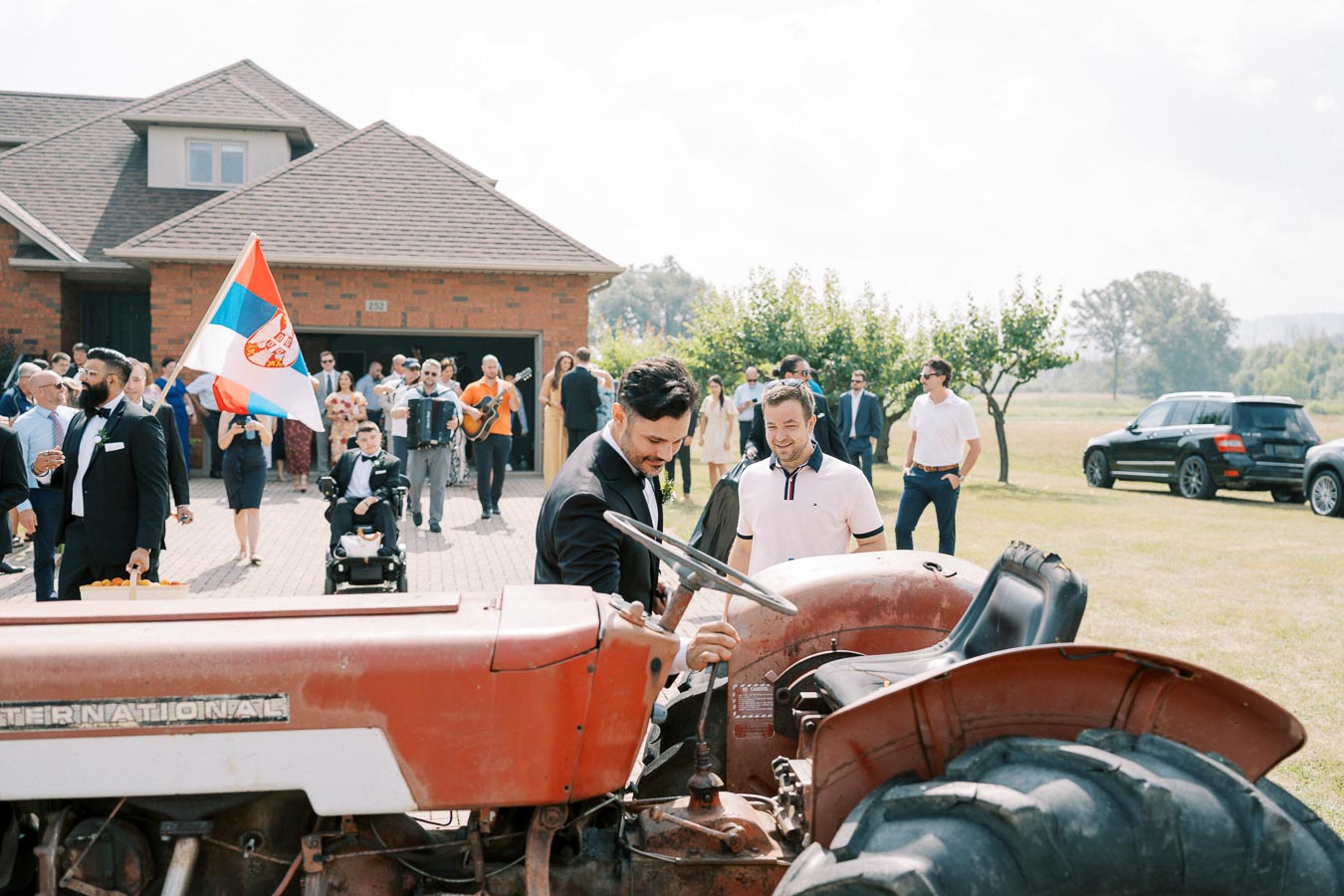 A group of well-dressed men gather around a vintage red tractor in front of a brick house. One man, holding a flag, leads the group while another interacts with the tractor. Musicians in the background create a festive atmosphere, and parked cars are seen in the grassy area.