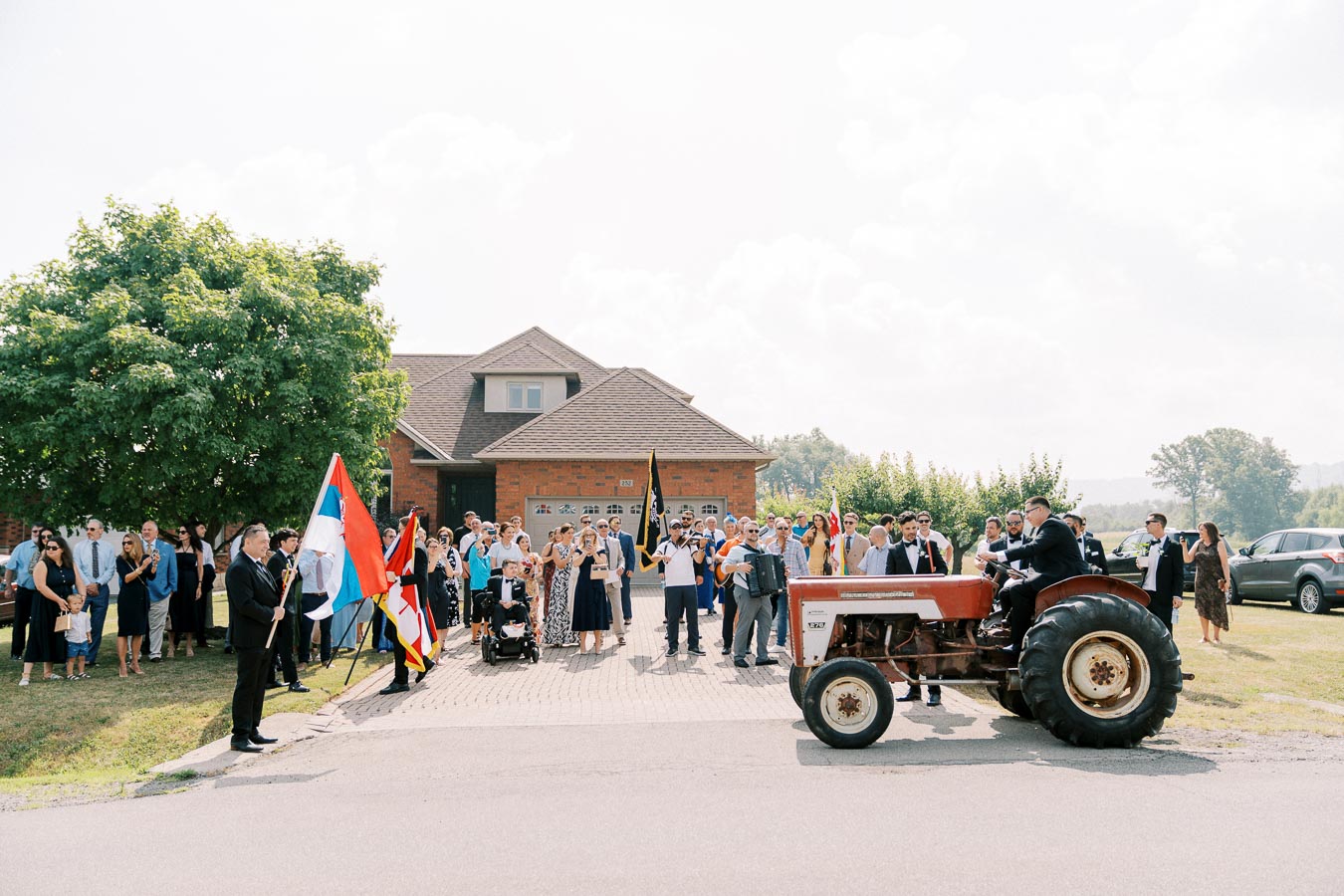 A group of people gathered in formal attire outside a suburban house, celebrating with flags near a red tractor.