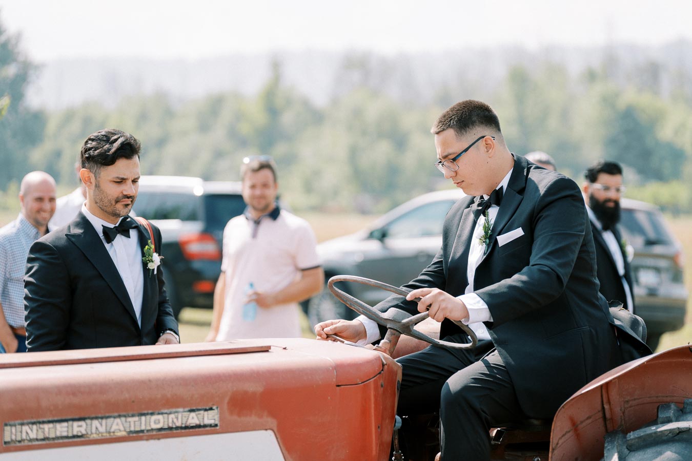 A man in a suit and bow tie sits on a vintage red tractor labeled International while another man in a similar suit stands nearby, surrounded by casually dressed people, outdoors in a sunny field with trees and cars in the background.