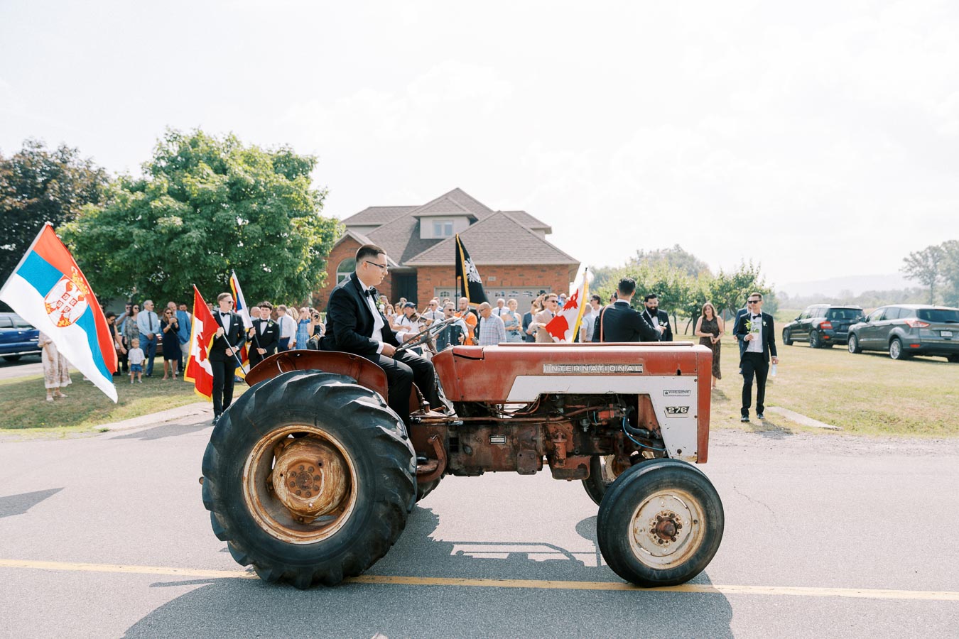 A group of well-dressed people in a parade featuring a red and white tractor, with flags and a crowd gathering on a sunny day.