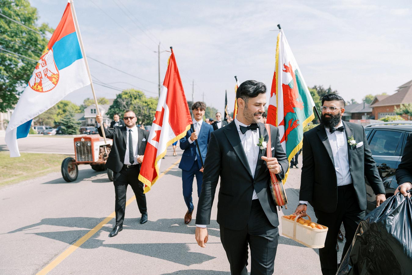 A group of men dressed in formal suits, carrying international flags, walk down a street during a wedding procession on a sunny day.