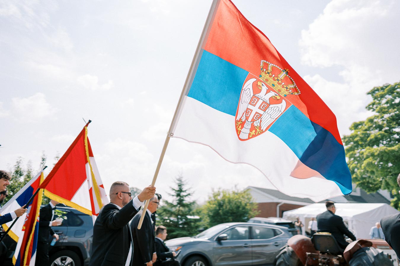 Man proudly waving Serbian flag during outdoor event with blue sky, trees, and cars in the background, celebrating national pride.