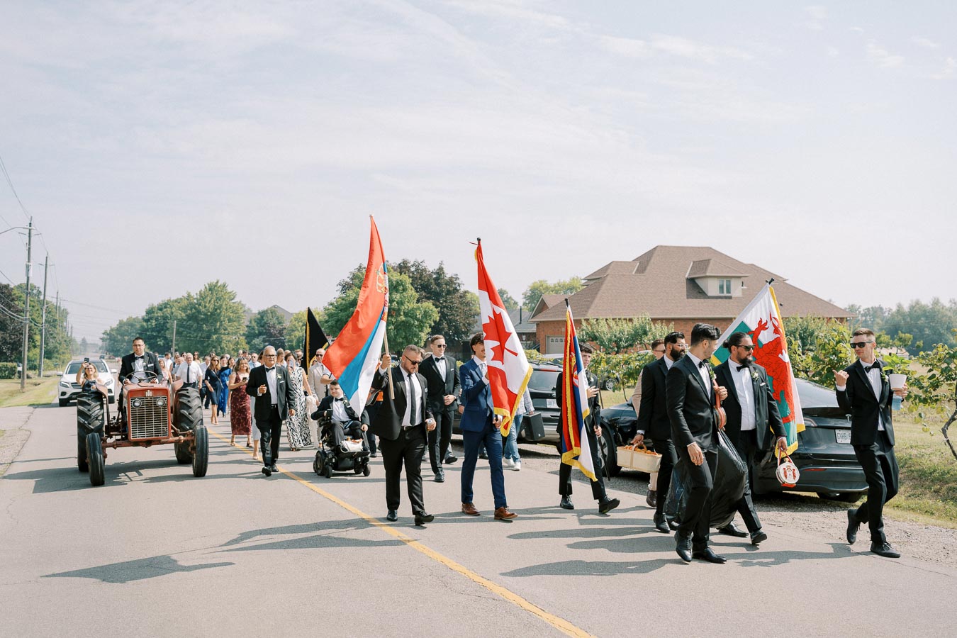 A procession of people, dressed in formal attire, walking along a suburban street. The group includes individuals carrying various flags, including the Canadian flag. A vintage tractor leads the parade with a suited driver, and onlookers follow in cars and along the sidewalk. A sunny day with clear skies and a residential backdrop sets the scene.