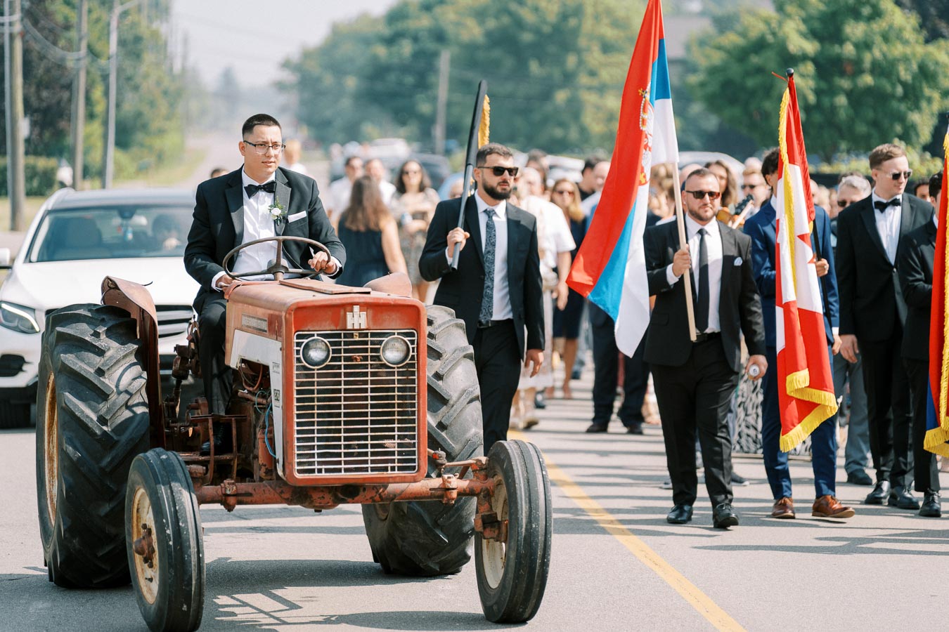 A man in a suit drives a vintage tractor in a parade, accompanied by several people holding flags and wearing formal attire. The event takes place on a sunny day with green trees in the background, blending tradition and celebration.