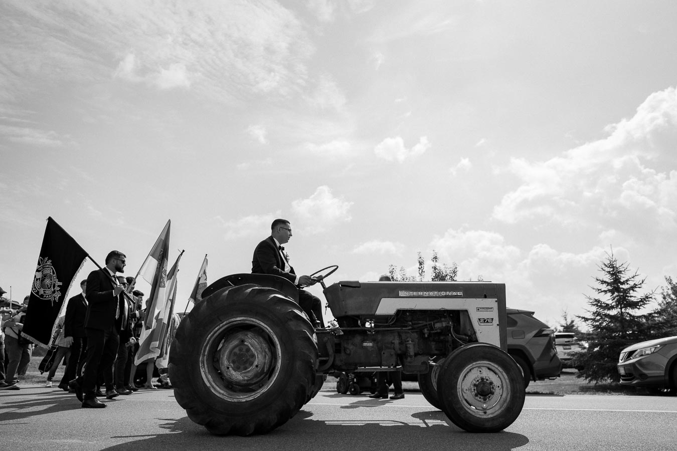 Black and white image of a man in a suit driving a vintage tractor in a parade, accompanied by a group of people carrying flags on a sunny day.