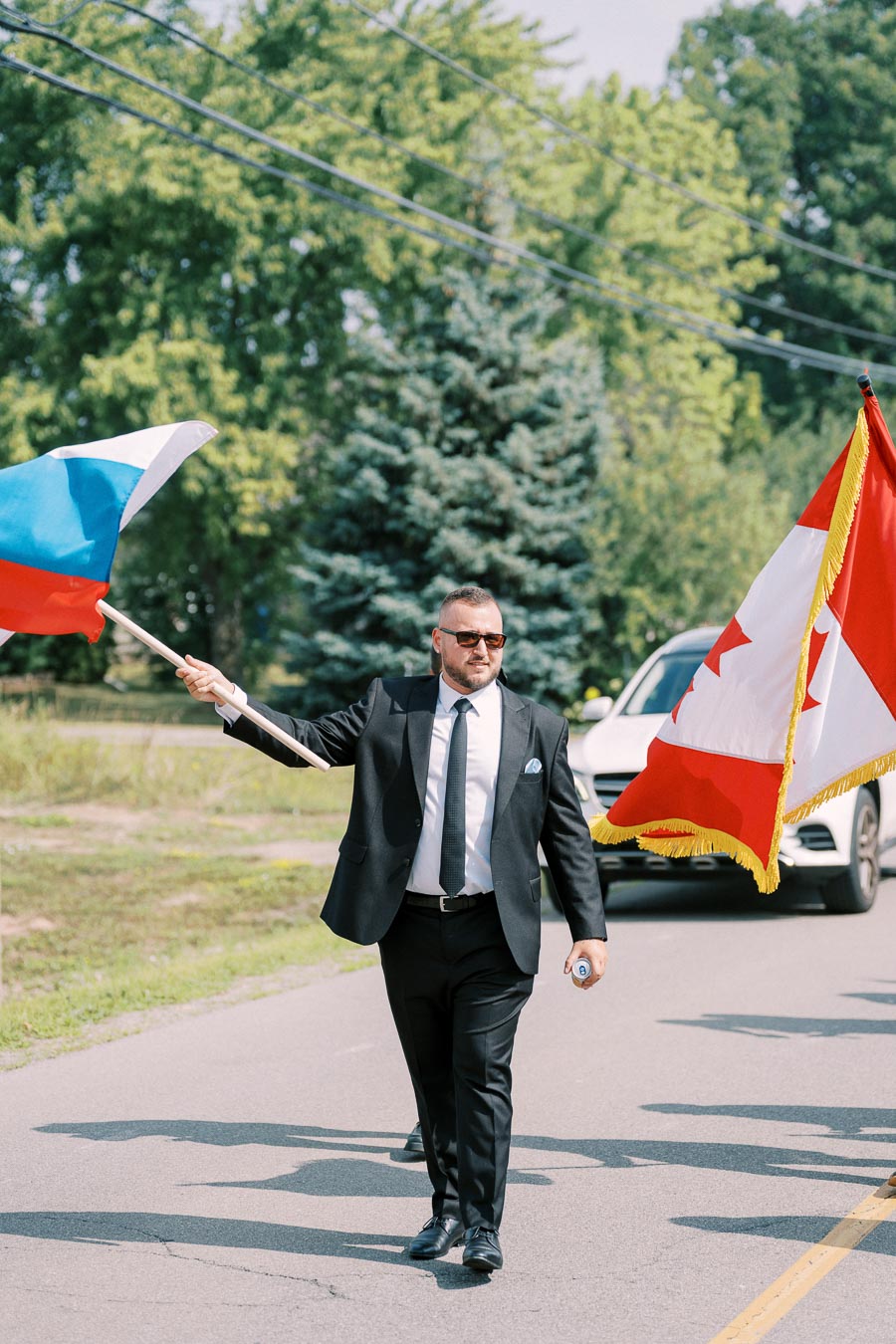 A man in a suit walking on a road while holding the flags of Russia and Canada, surrounded by greenery and a car in the background.