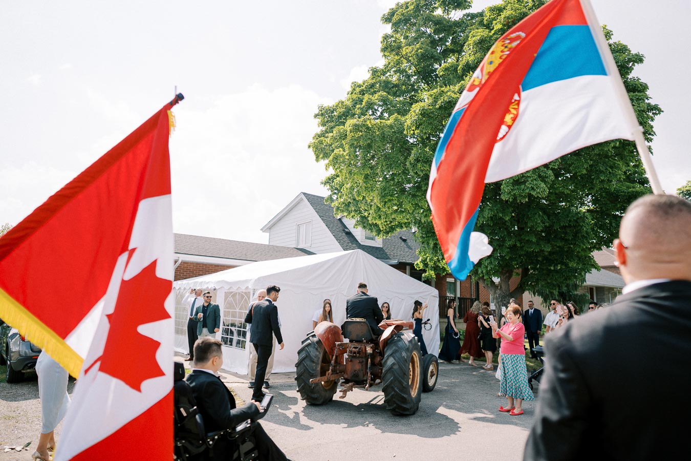 Multicultural event with Canadian and Serbian flags, people in formal attire gathered around a tractor in front of a white tent, suggesting a festive gathering in Canada.