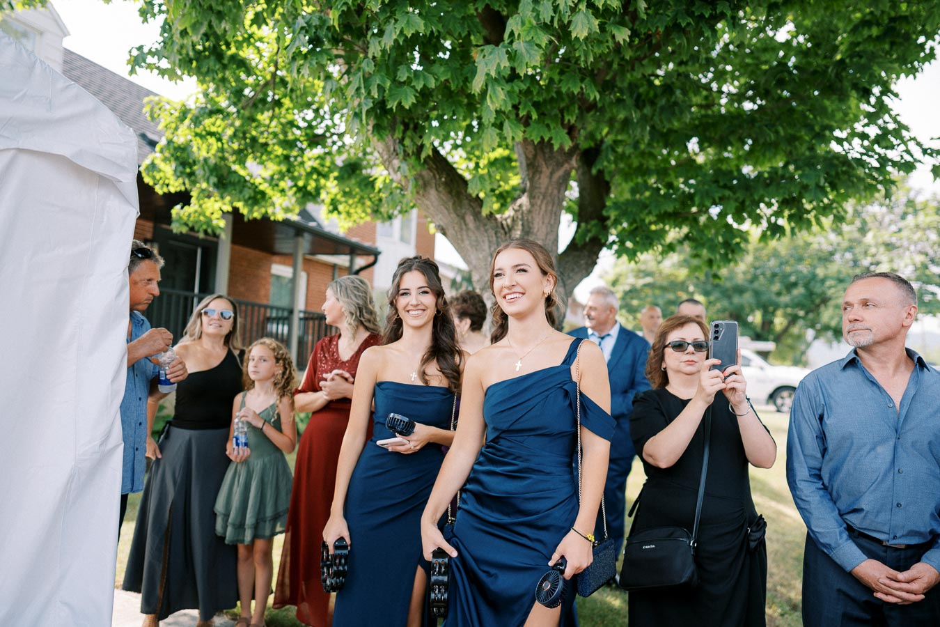 A group of people attending an outdoor event, with two women in elegant blue dresses standing at the front, smiling. The scene is set under a large tree, with attendees dressed in formal and semi-formal attire.