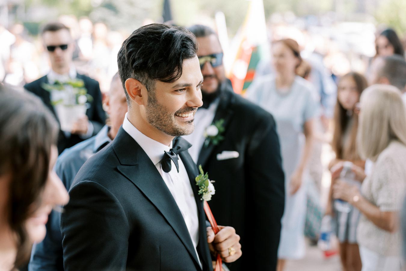 A groom in a black tuxedo smiling during an outdoor wedding ceremony with guests in the background.