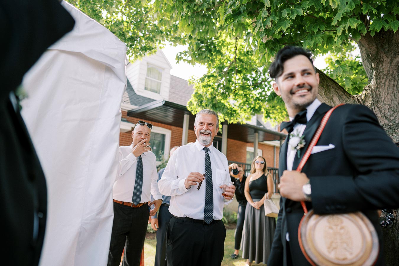 A joyful outdoor wedding celebration with well-dressed guests, including a smiling man in a tuxedo holding an ornate wooden item, surrounded by people in formal attire beneath a green tree.