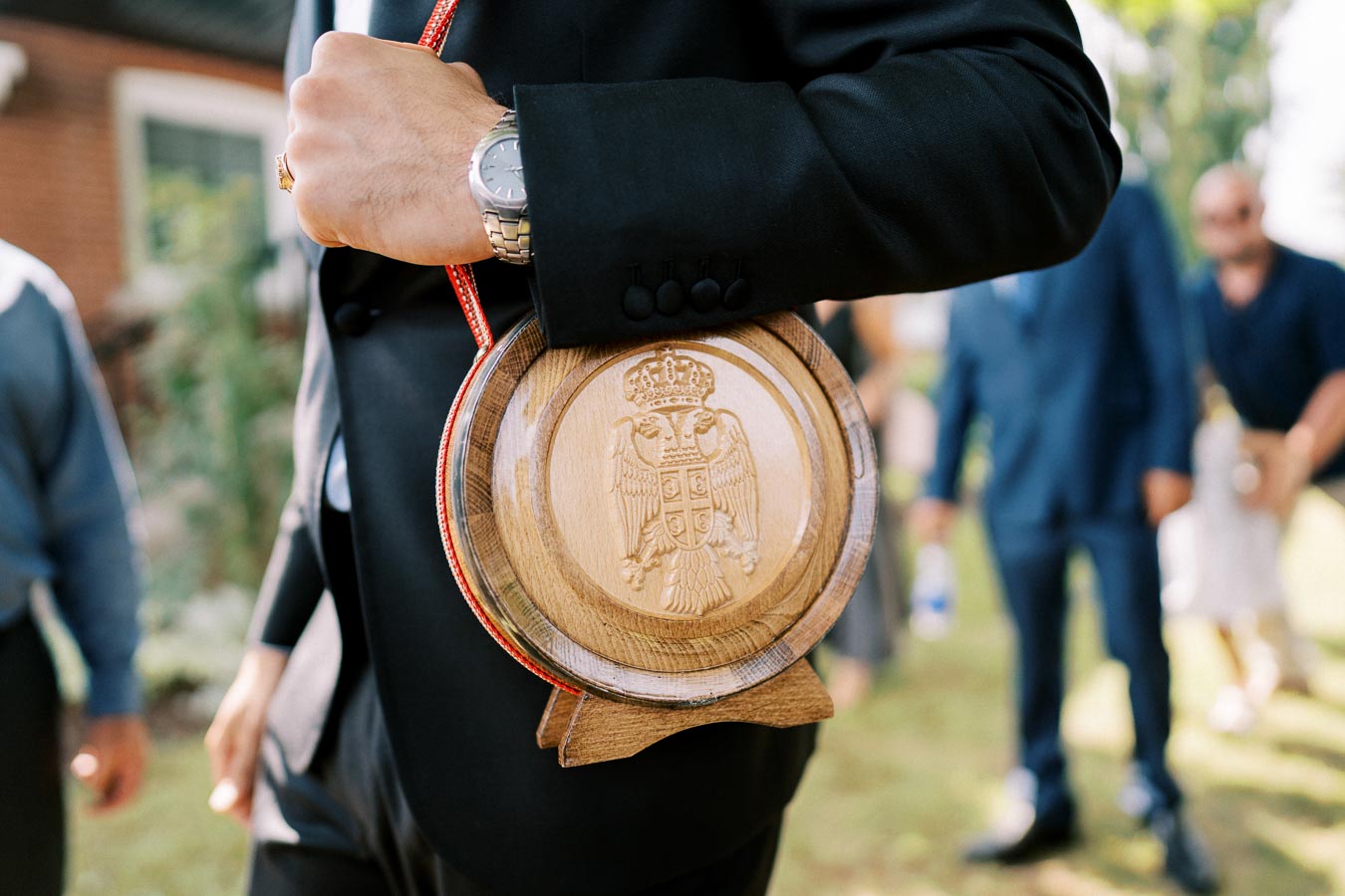 A man in a suit holds a traditional wooden flask with a carved emblem, standing outdoors at an event or ceremony.